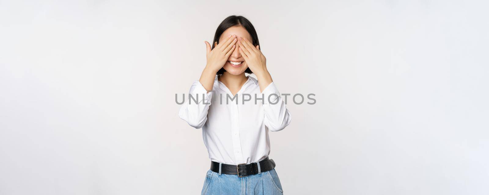 Image of young asian woman close her eyes and smiles, waits for surprise, anticipates, stands over white background.