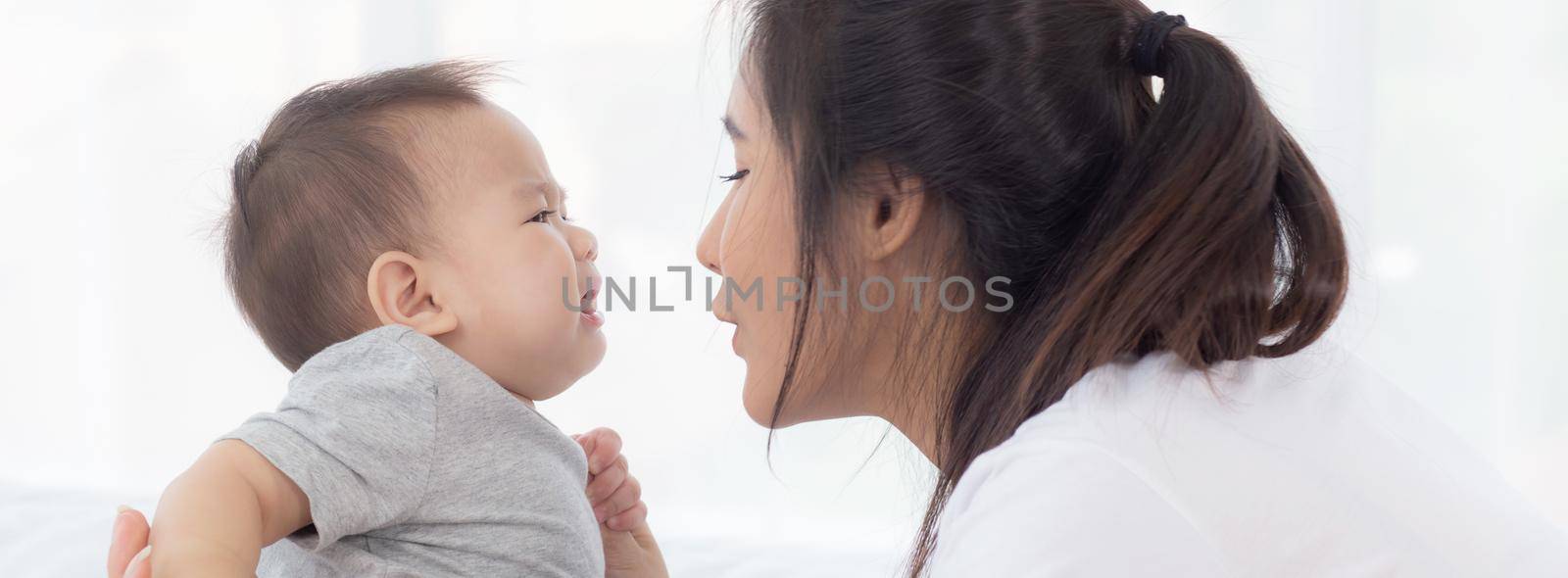 Young asian mother holding little baby girl together in the bedroom at home, mom carry child and care, woman and kid with carefree, toddler and parent, emotion and expression, family concept. by nnudoo