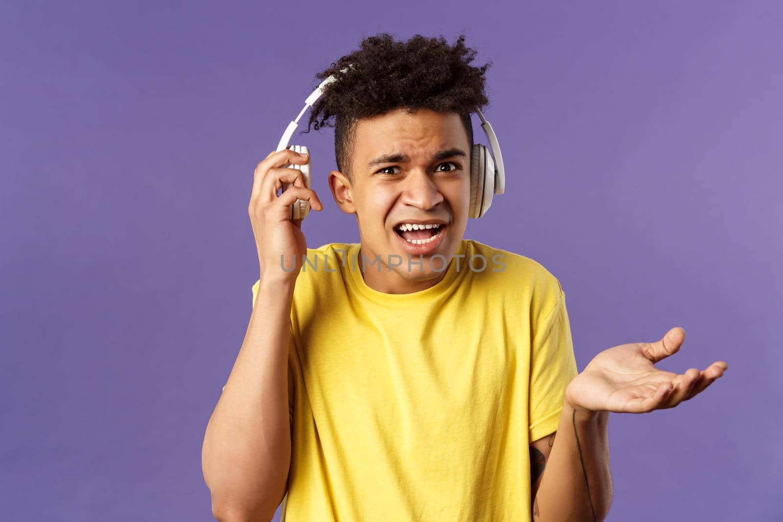 What do you want, I am in headphones. Portrait of confused annoyed young man shrugging, take-off earphone to hear what person asked while he was listening music, purple background.