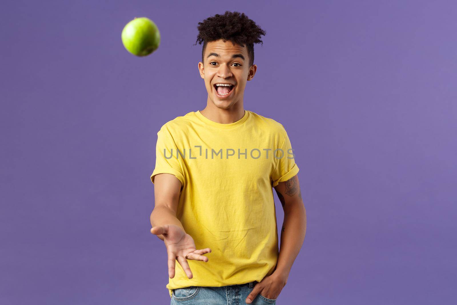 Holidays, vitamins and vacation concept. Portrait of handsome upbeat young male student asking friend something eat, catching apple and smiling happy, standing purple background by Benzoix