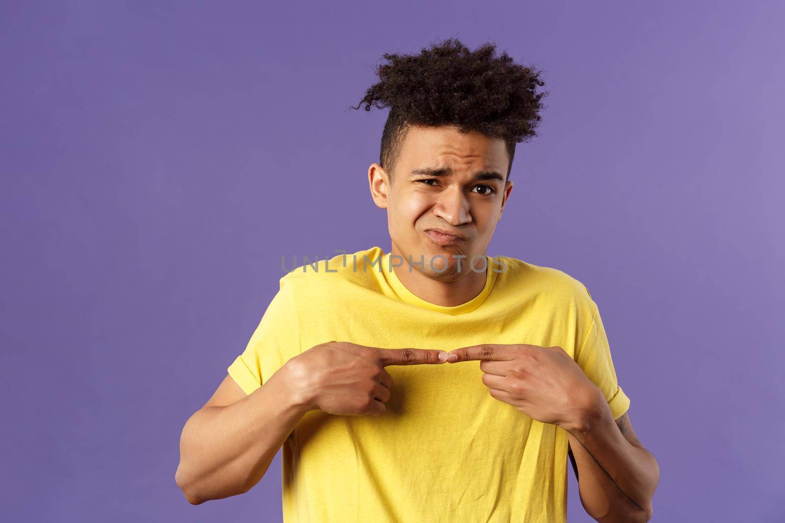 Close-up portrait of shy and modest young silly hispanic man trying say something but being too insecure, grimacing and frowning, look timid, two fingers touching pose, purple background.