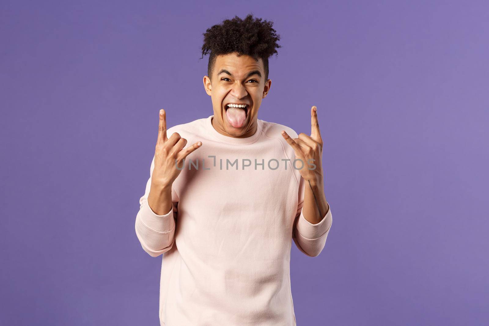 Portrait of enthusiastic, excited funny young man having fun, enjoying awesome rock concert, show heavy metal sign, stick tongue and rejoicing hear favorite band on stage, purple background.