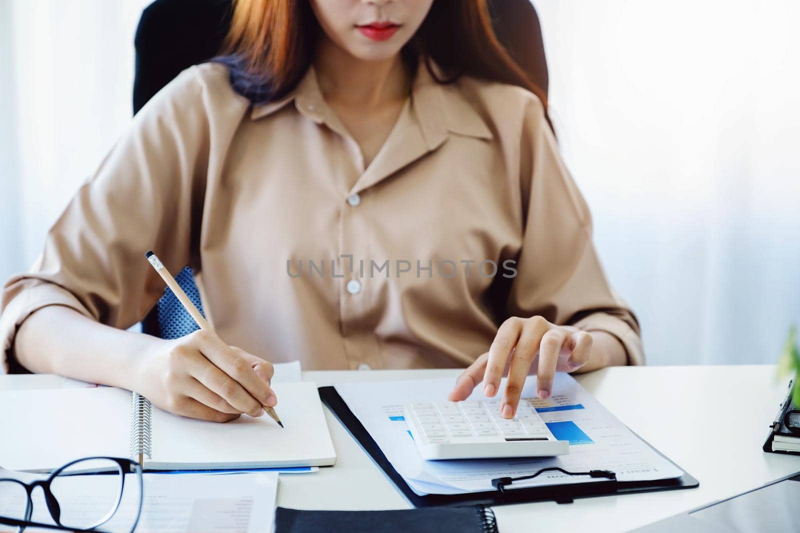 Financial and investment auditing concept, female auditor is pressing calculator to check budget and correctness of company profits through paperwork to calculate annual tax. by Manastrong