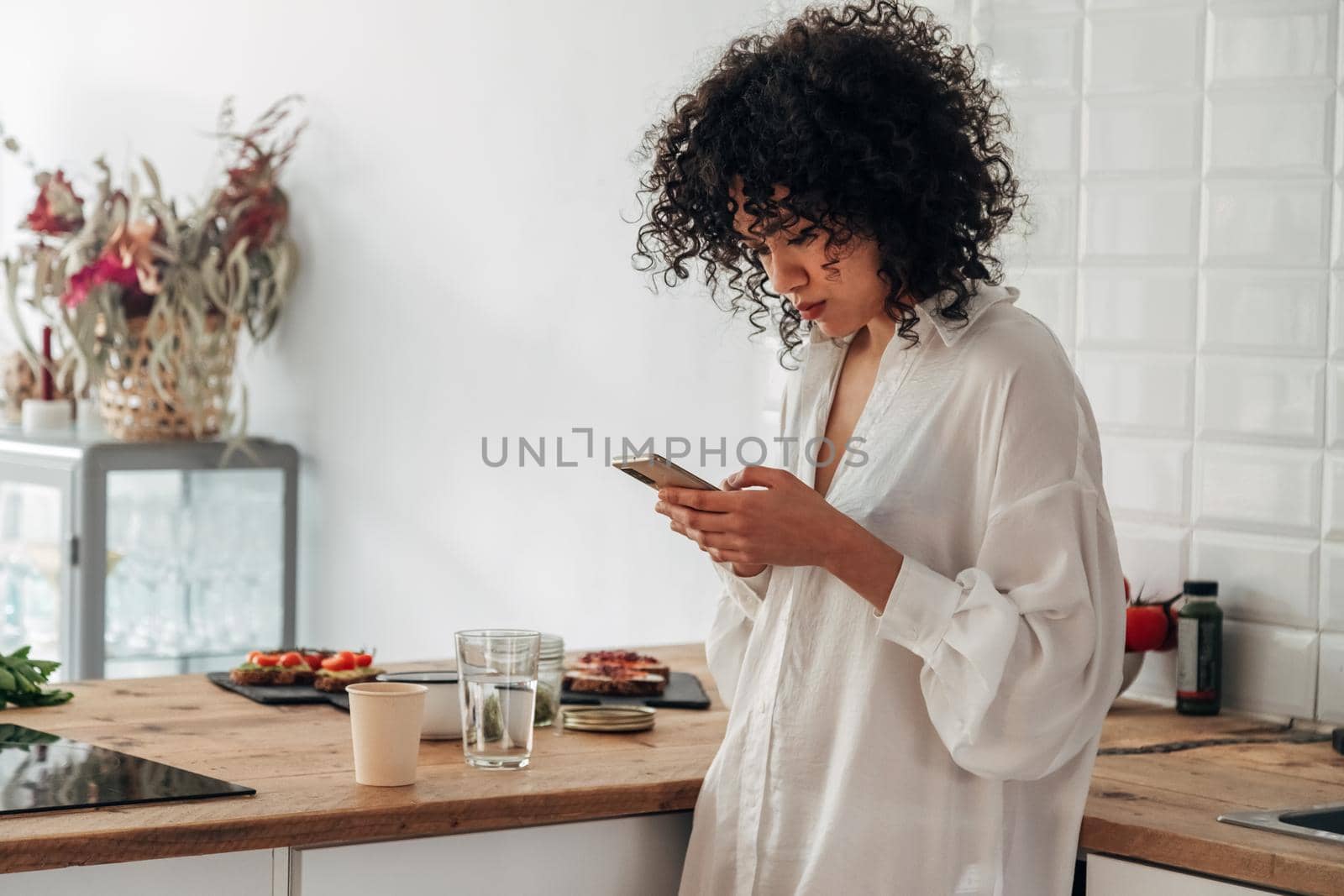 Young mixed race woman using mobile phone to send text message at home kitchen. Copy space. by Hoverstock