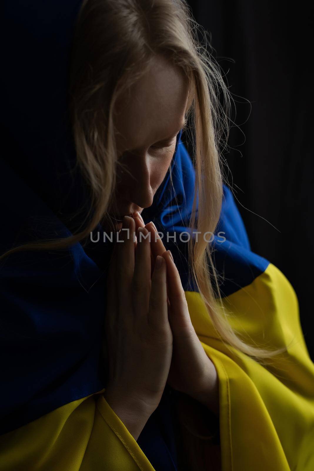 Ukrainian blonde woman prays with closed eyes for Ukraine during the war with the state flag on her head. Russia attacked Ukraine on February 24, 2022