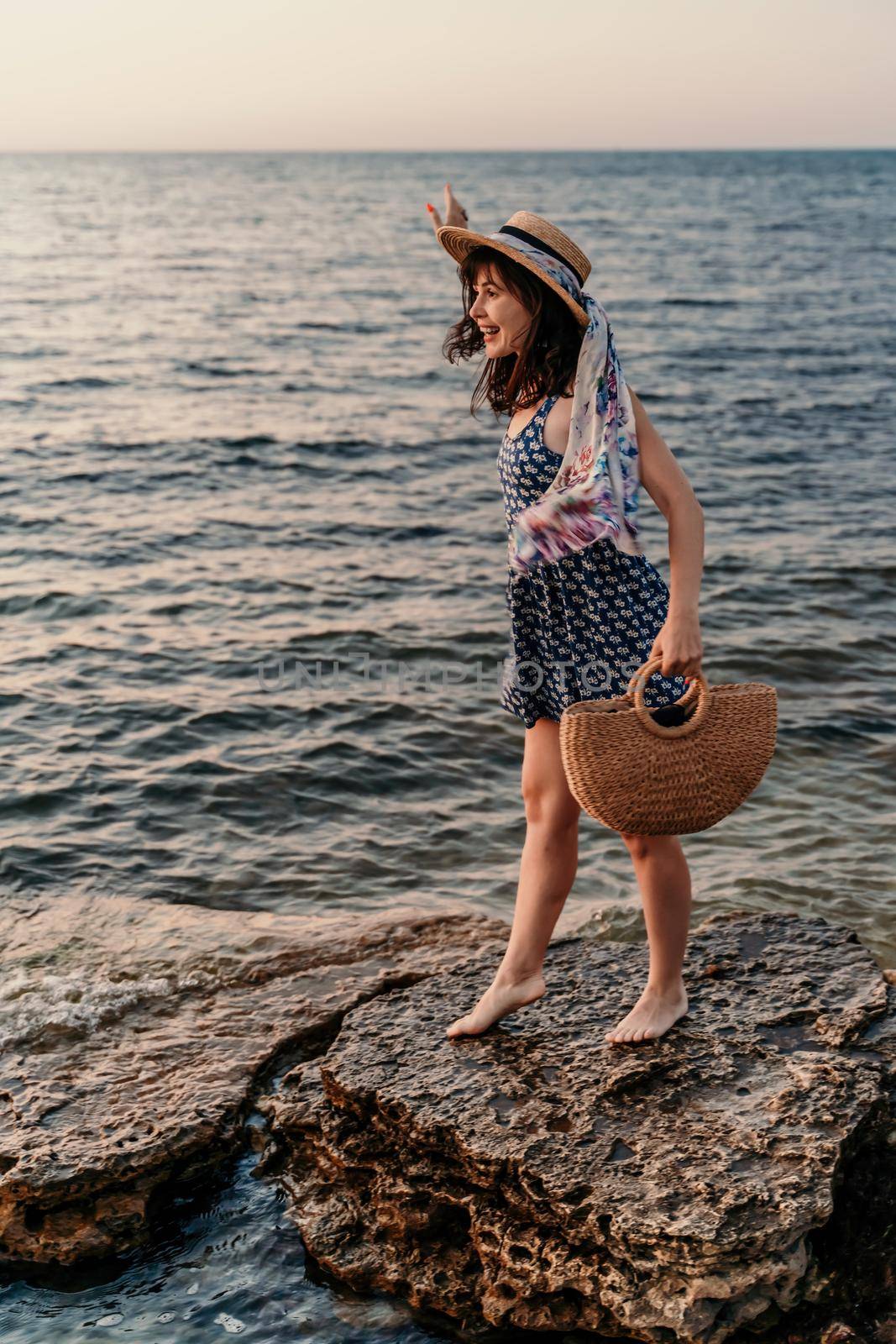 A woman in a dress, hat and with a straw bag is standing on the beach enjoying the sea. Happy summer holidays.