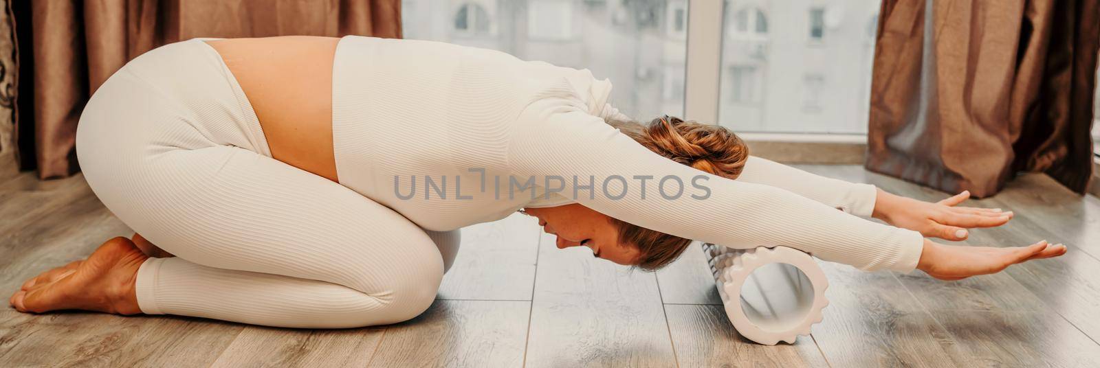 Adult athletic woman, in white bodysuit, performing fascia exercises on the floor - caucasian woman using a massage foam roller - a tool to relieve tension in the back and relieve muscle pain - the concept of physiotherapy and stretching training by Matiunina