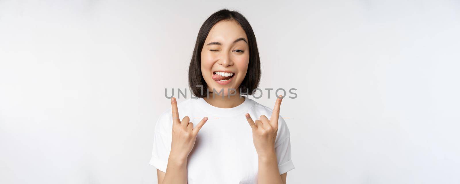 Sassy asian girl shouting, enjoying concert or festival, showing rock on, heavy metal sign, having fun, standing over white background.