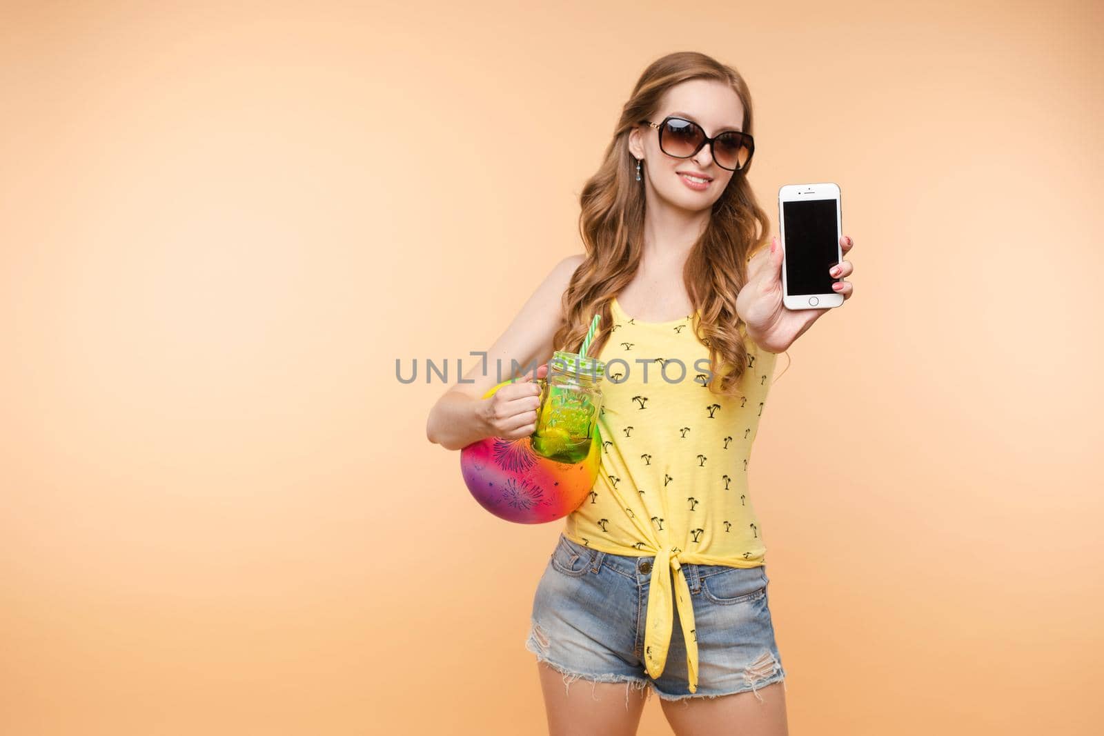 A woman in bright clothes, a yellow T-shirt and denim shorts, sunglasses holds a ball and cold lemonade in her hands, on an isolated background.