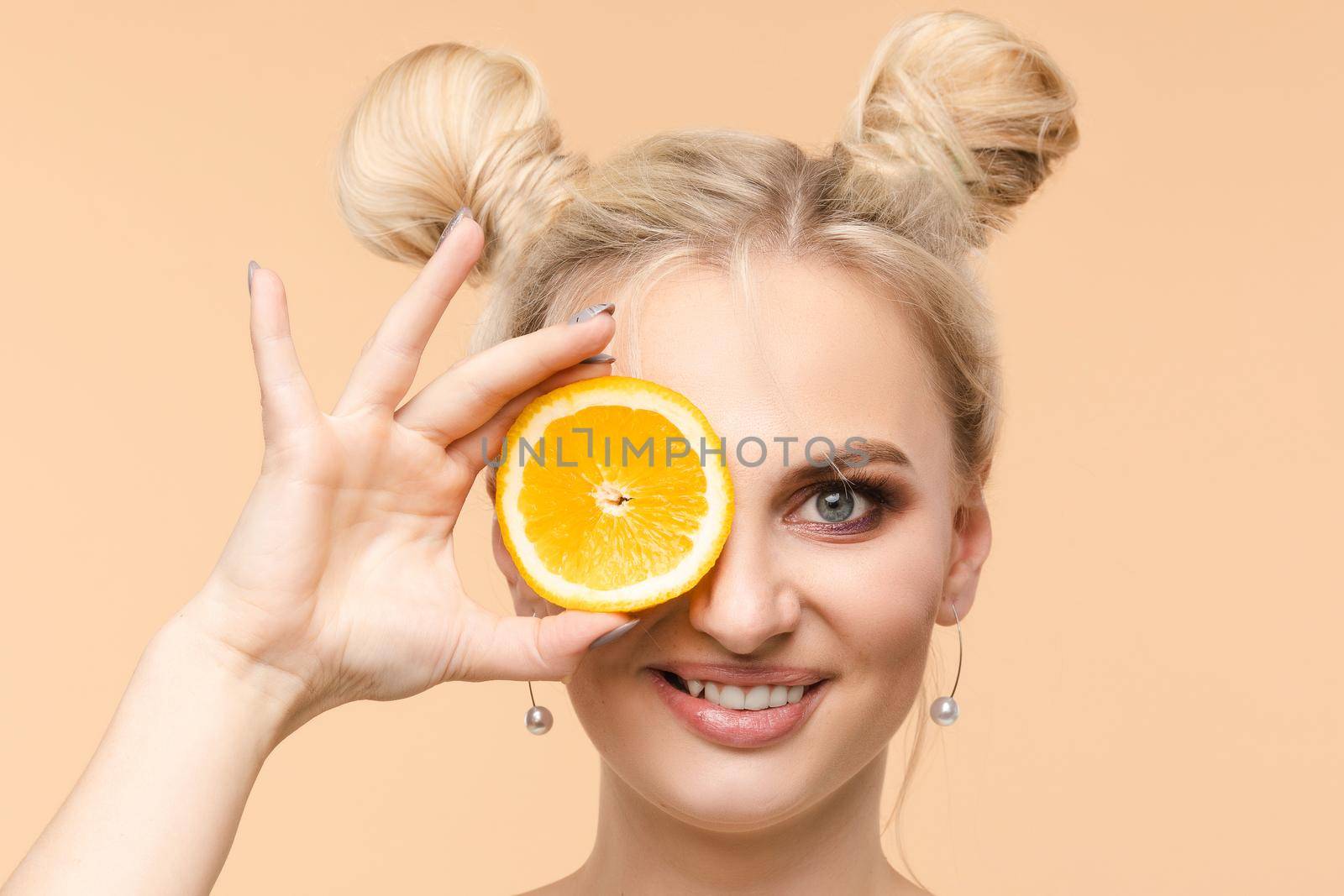 Pretty charming joyful attractive cheerful funny comic positive nude natural pure girl having two pieces of orange, closing one eye, isolated on beige background with copy space for advertisement