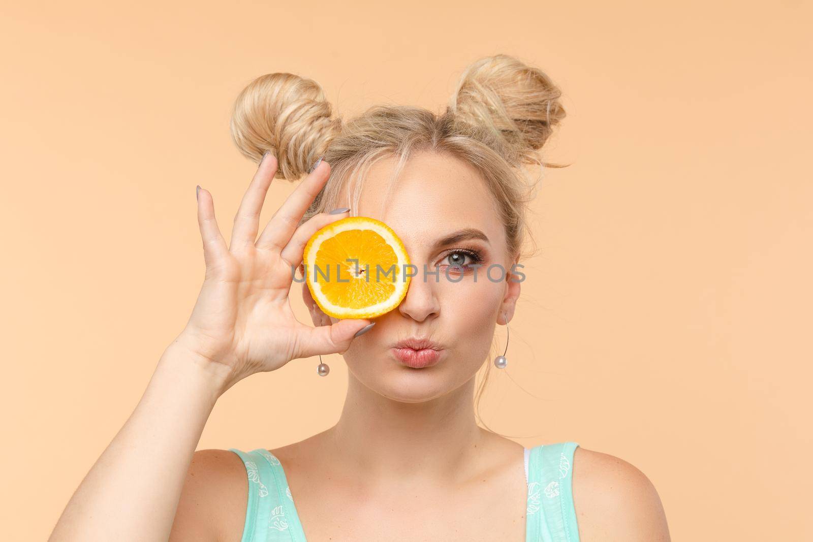 Pretty charming joyful attractive cheerful funny comic positive nude natural pure girl having two pieces of orange, closing one eye, isolated on beige background with copy space for advertisement