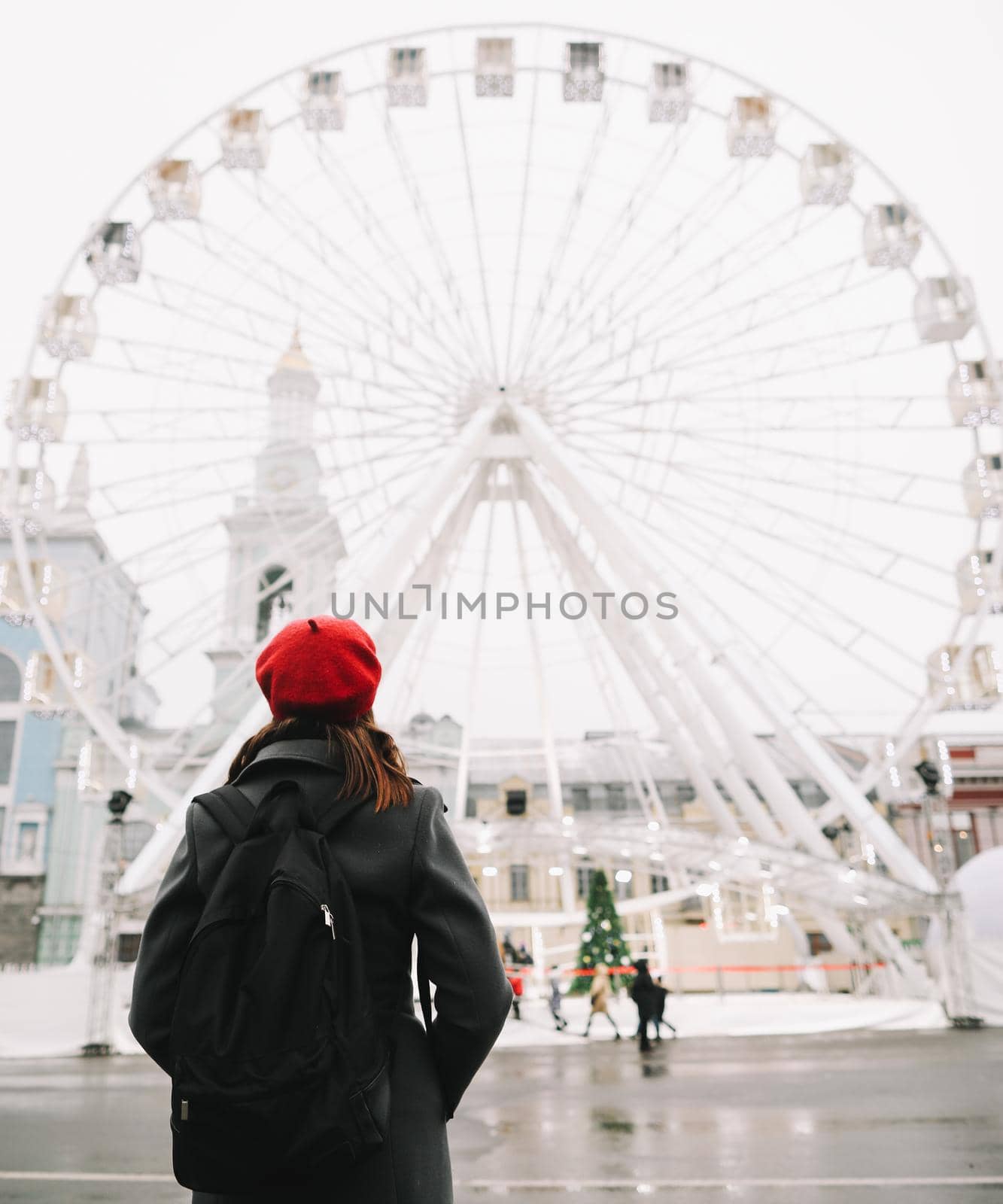 Young woman traveler in red hat and black coat looking at the ferris wheel in Kyiv, Ukraine. Outdoors photo of a girl on the background of modern city architecture.