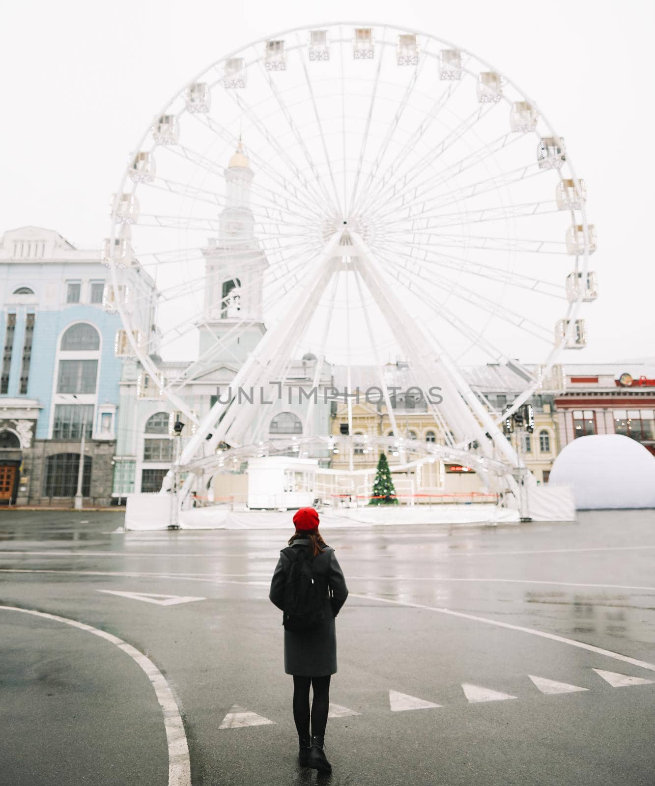 Young woman traveler in red hat and black coat looking at the ferris wheel in Kyiv, Ukraine. Outdoors photo of a girl on the background of modern city architecture.