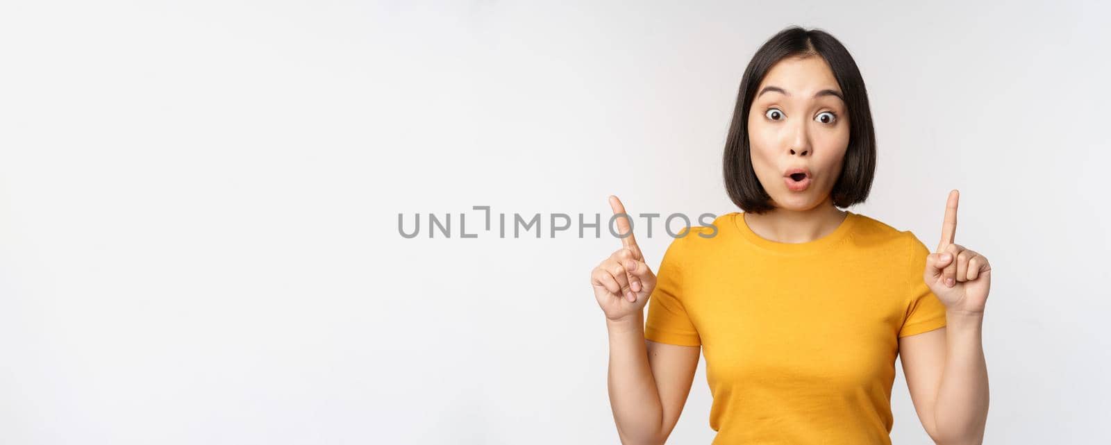 Portrait of impressed asian girl, pointing fingers up, say wow, looking amazed at camera, standing over white background. Copy space