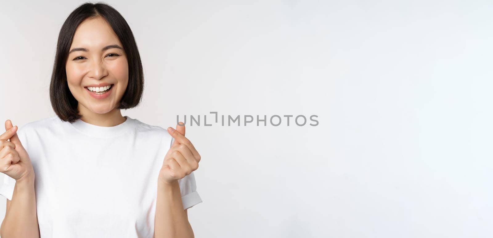 Beautiful asian woman smiling, showing finger hearts gesture, wearing tshirt, standing against white background by Benzoix