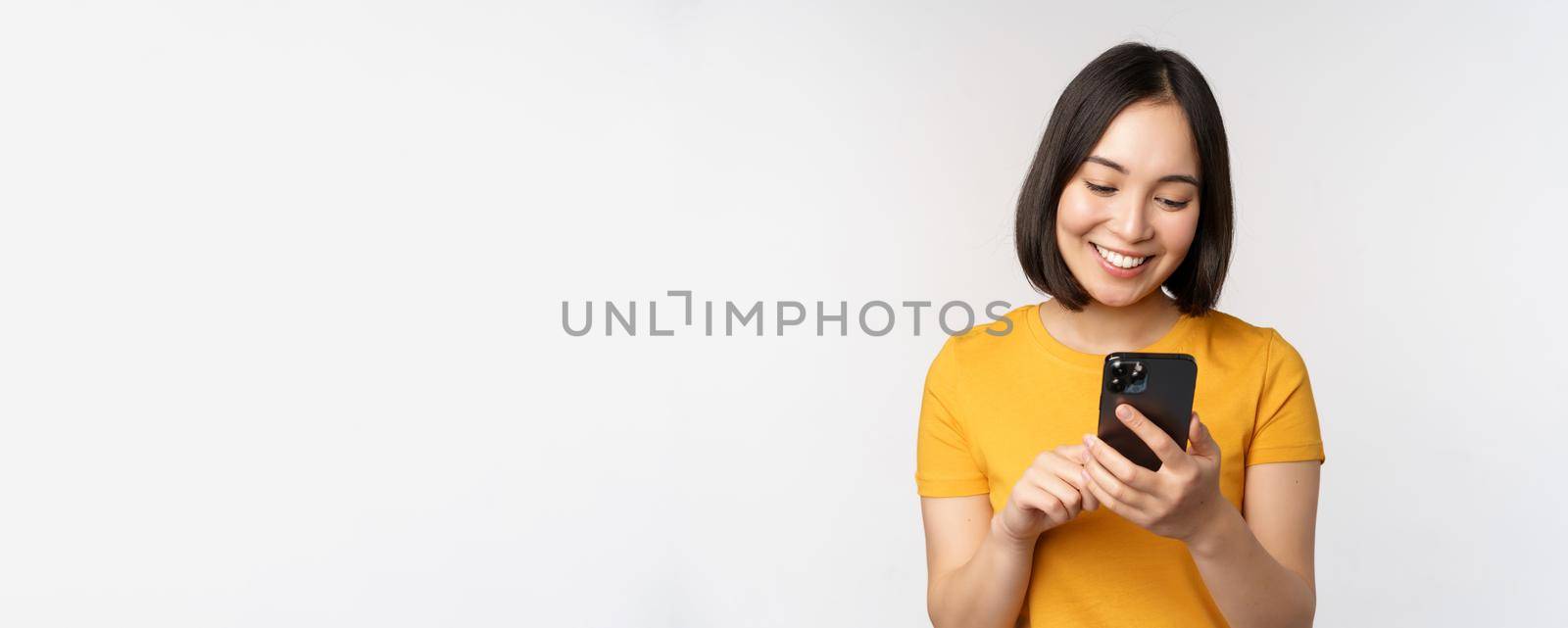 People and technology concept. Smiling asian girl using smartphone, texting on mobile phone, standing against white background by Benzoix