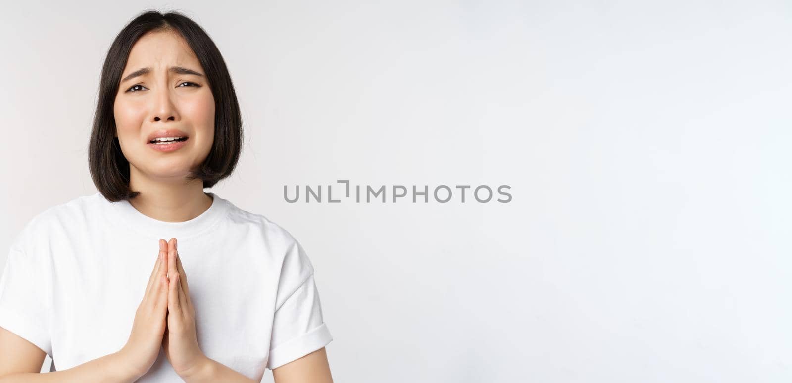 Desperate crying asian woman begging, asking for help, pleading and say please, standing in white t-shirt over white background by Benzoix