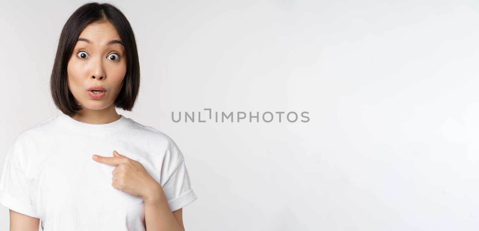 Portrait of surprised asian girl student, pointing finger left, raising eyebrows amazed, impressed by big deal, offer ahead, standing over white background by Benzoix