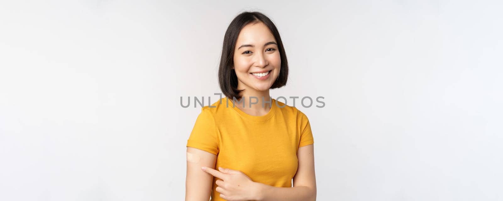 Covid-19 vaccination campaign. Smiling asian girl pointing at her band aid on shoulder, recommending get vaccine from omicron coronavirus, white background by Benzoix