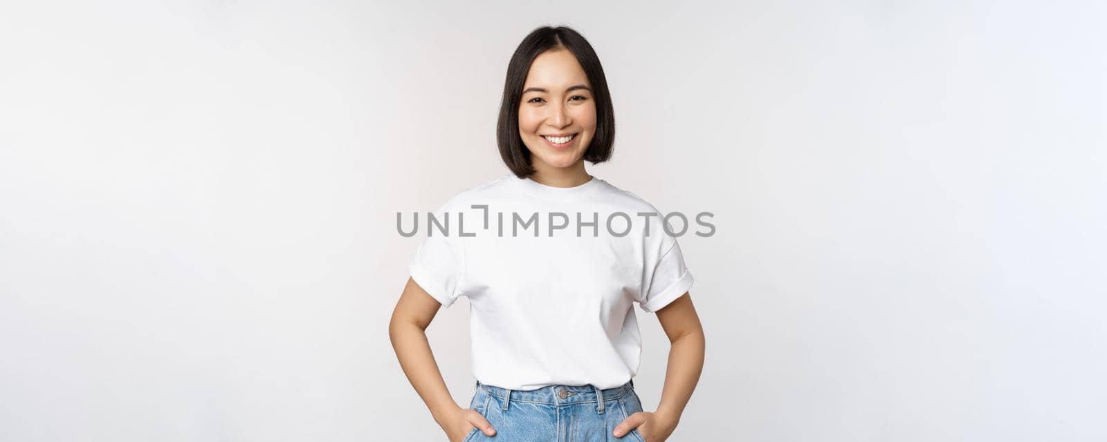 Lifestyle. Happy modern asian girl, smiling and looking happy at camera, posing in white tshirt and jeans, studio background.