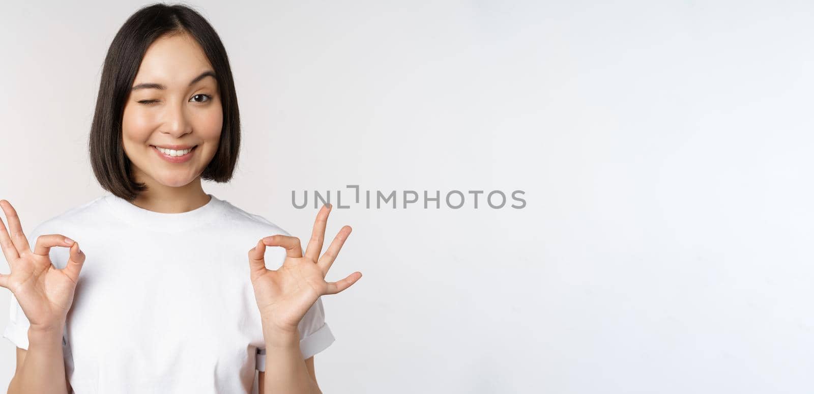 Very well, excellent. Smiling asian woman showing okay sign, approval, ok gesture, looking satisfied, recommending smth, standing over white background by Benzoix