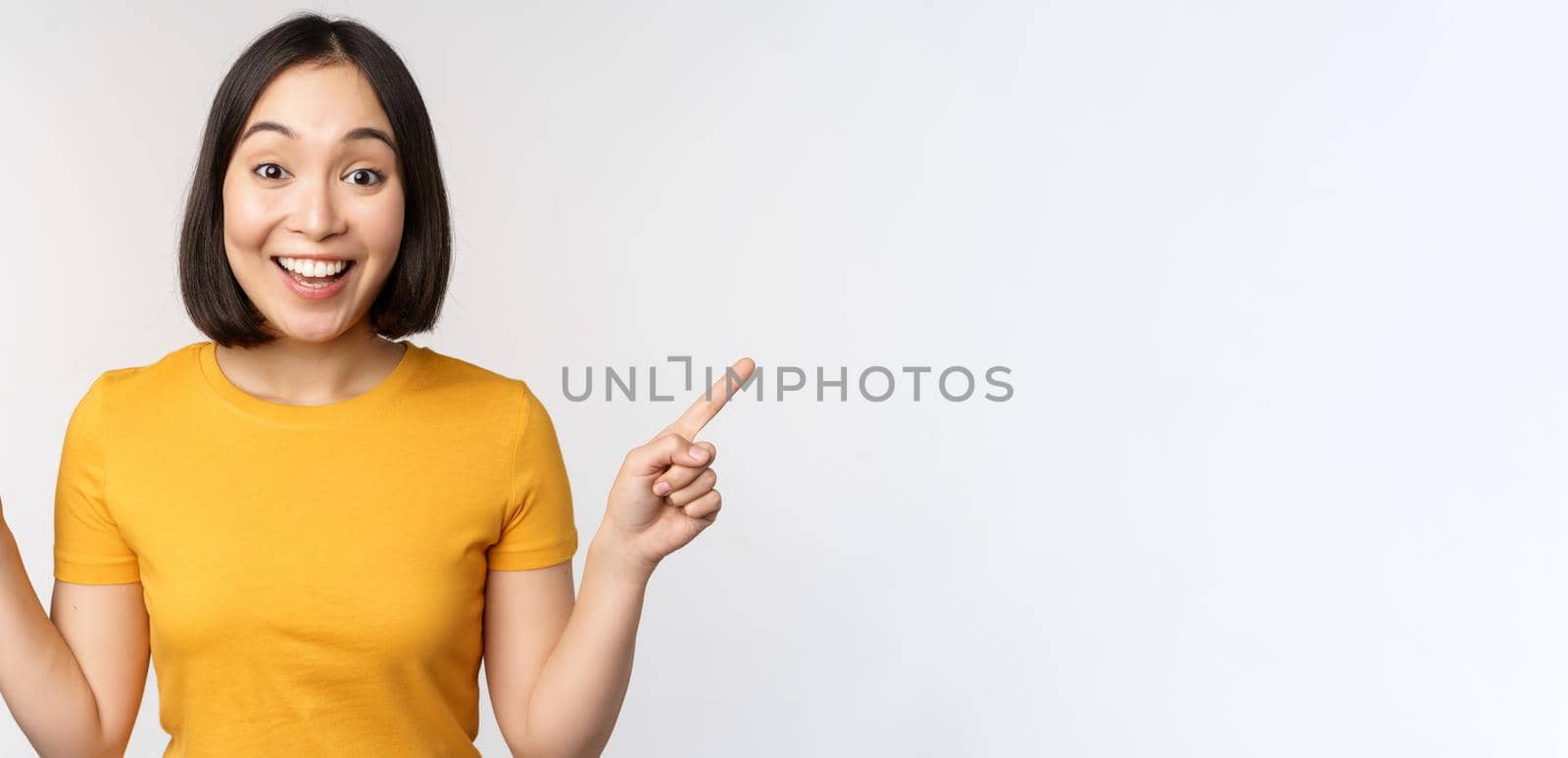 Cute asian girl pointing fingers sideways, showing left and right promo, two choices, variants of products, standing in yellow tshirt over white background.