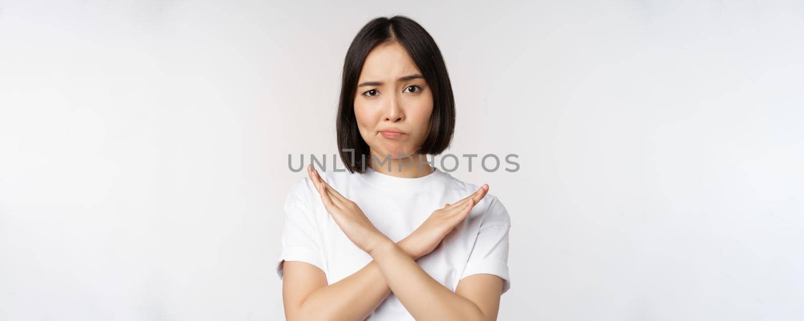 Portrait of asian korean woman showing stop, prohibition gesture, showing arm cross sign, standing in tshirt over white background. Copy space