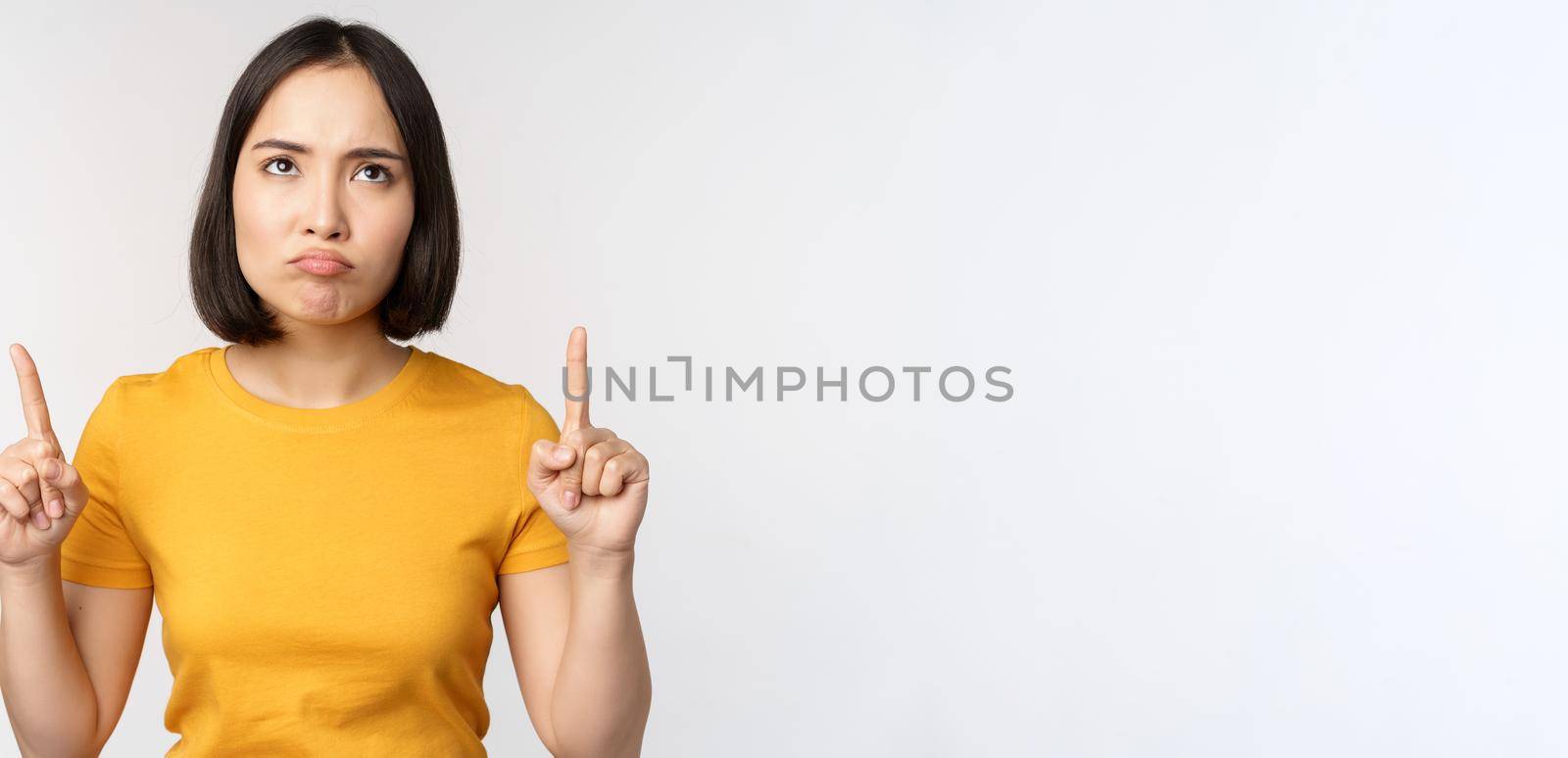 Disappointed asian woman looking, pointing fingers up with angry moody face expression, standing in yellow tshirt over white background by Benzoix