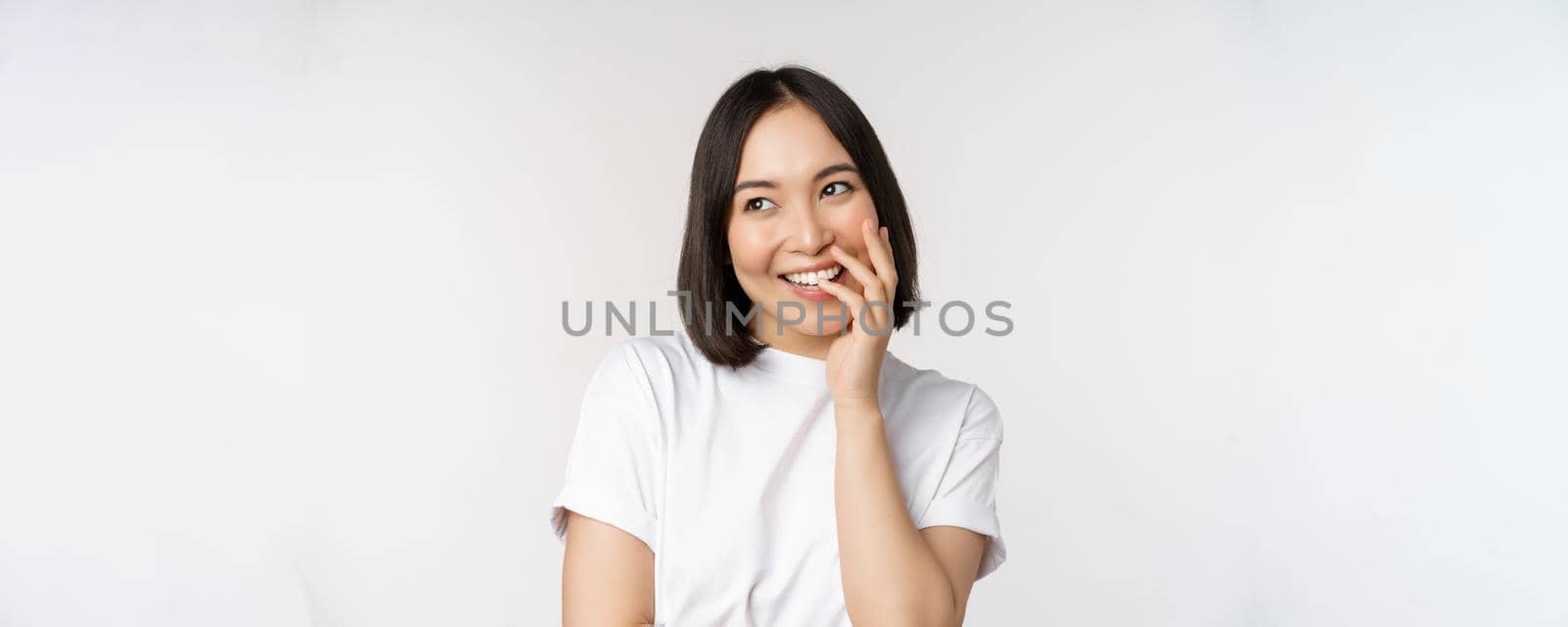 Portrait of young beautiful woman, korean girl laughing and smiling, looking coquettish, standing against white background.