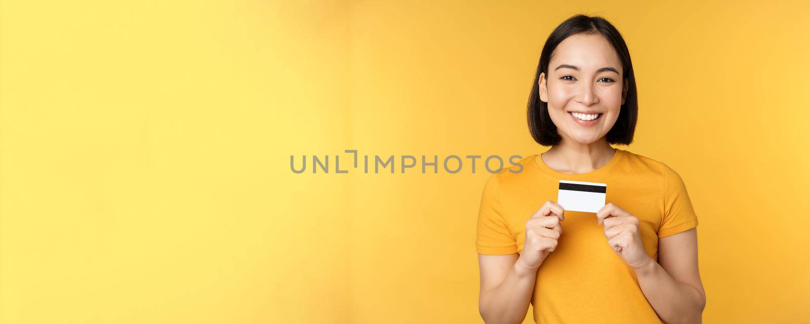 Beautiful asian woman showing credit card and smiling, recommending bank service, standing over yellow background by Benzoix