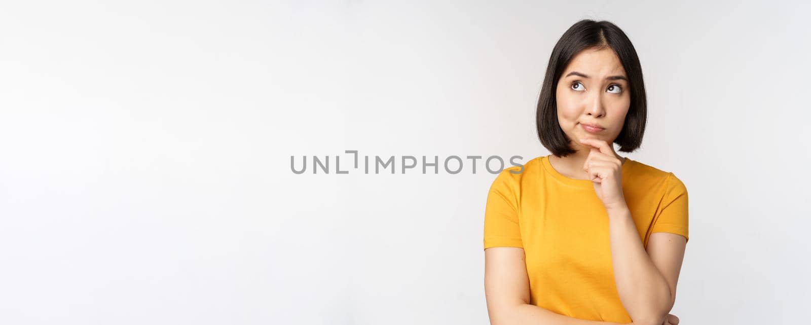 Image of thinking asian woman, looking aside and pondering, making decision, standing in yellow tshirt over white background by Benzoix