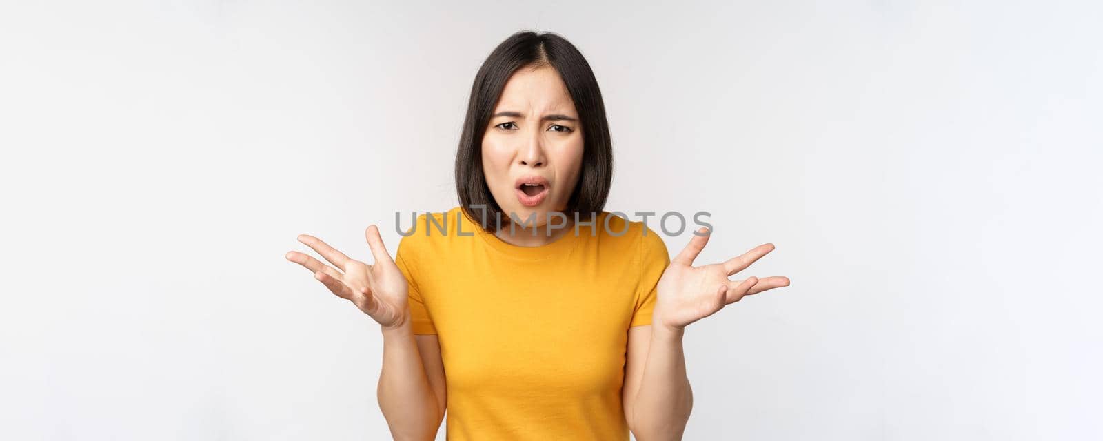 Frustrated, confused asian woman cant understand smth, looking clueless, shrugging shoulders, standing in yellow tshirt over white background by Benzoix