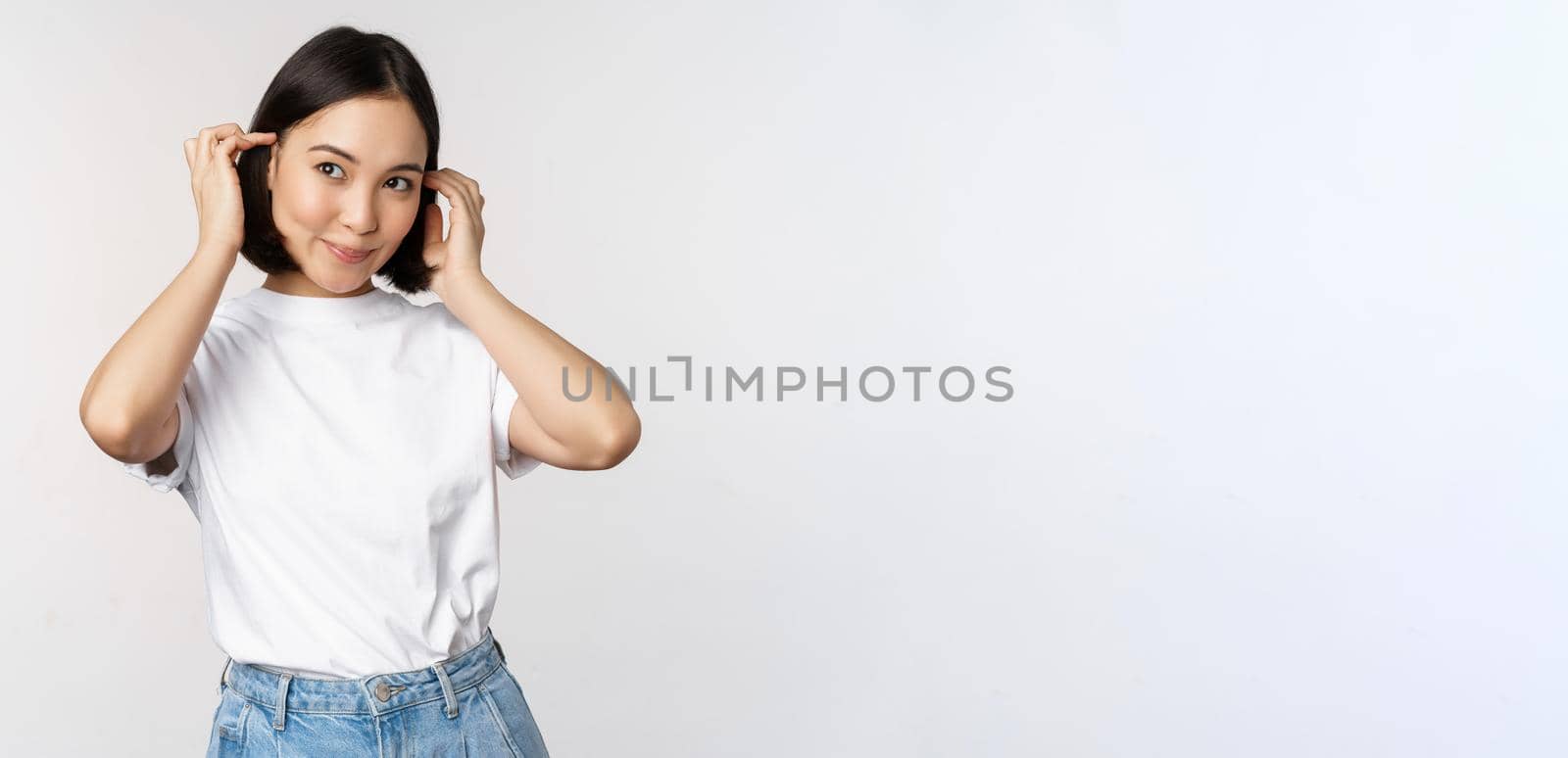 Portrait of cute, beautiful asian woman touching her new short haircut, showing hairstyle, smiling happy at camera, standing over white background by Benzoix