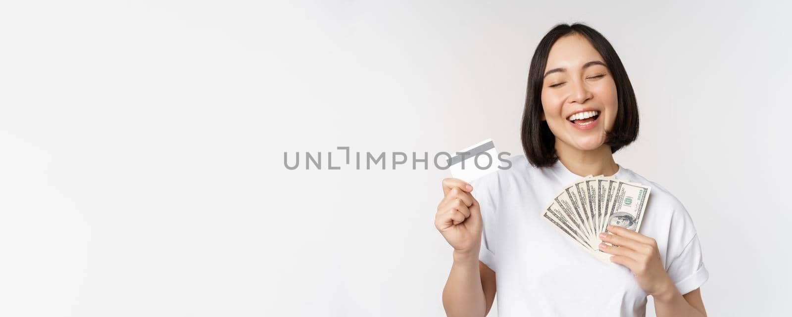 Portrait of asian woman smiling, holding credit card and money cash, dollars, standing in tshirt over white background.