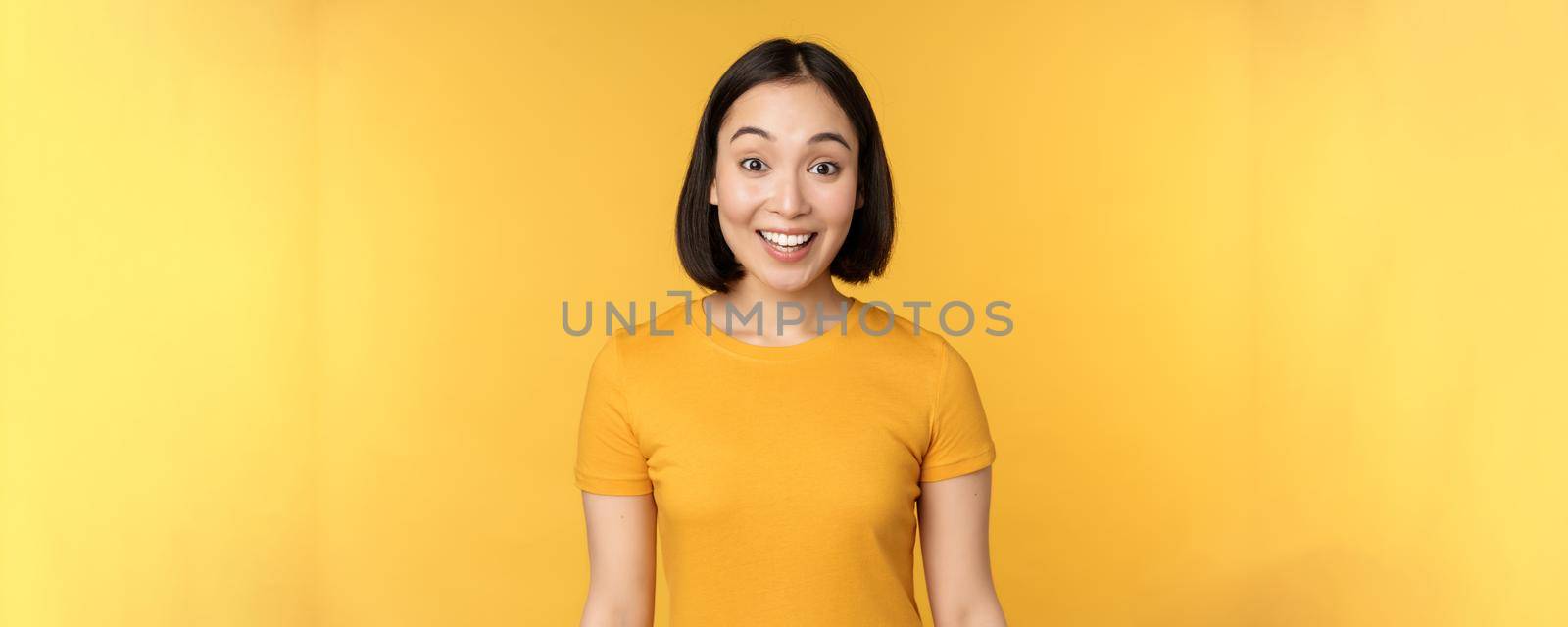 Image of asian girl looking surprised, reacting amazed, raising eyebrows impressed, standing over yellow background by Benzoix