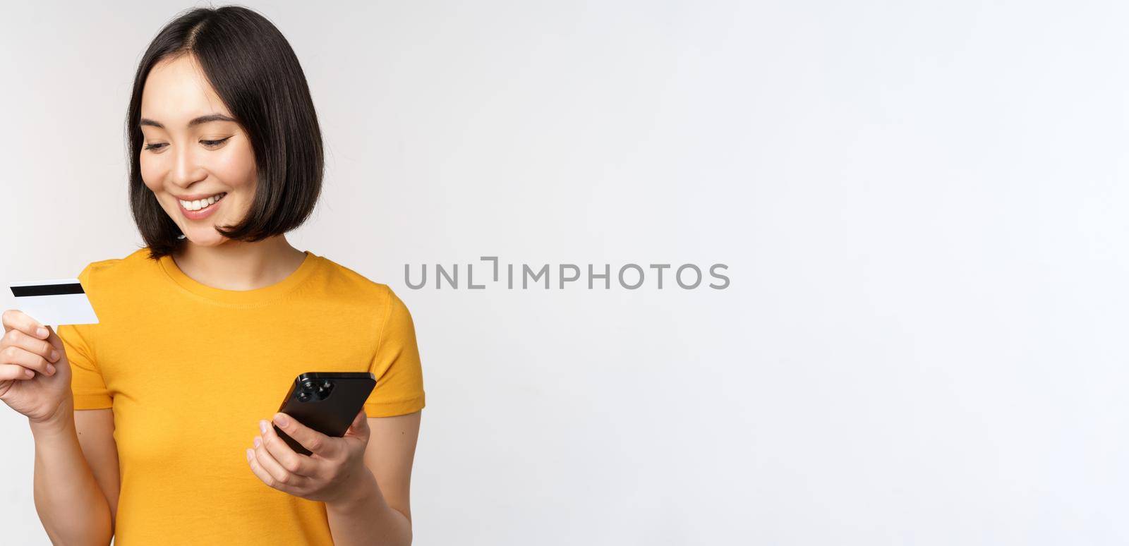 Beautiful smiling asian girl using credit card and mobile phone, paying online on smartphone, standing in yellow tshirt over white background.