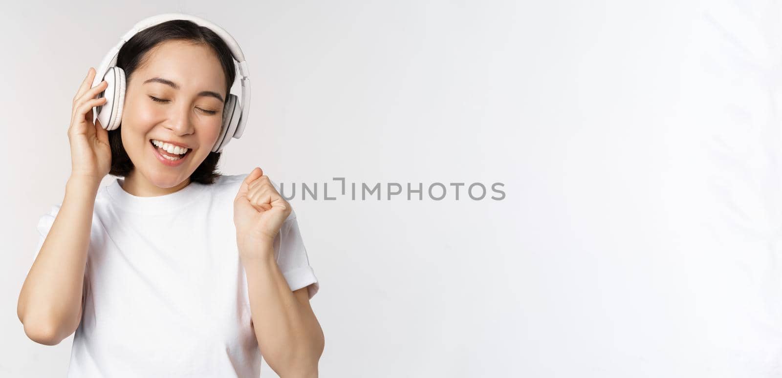 Modern asian girl dancing, listening music with headphones, smiling happy, standing in tshirt over white background by Benzoix