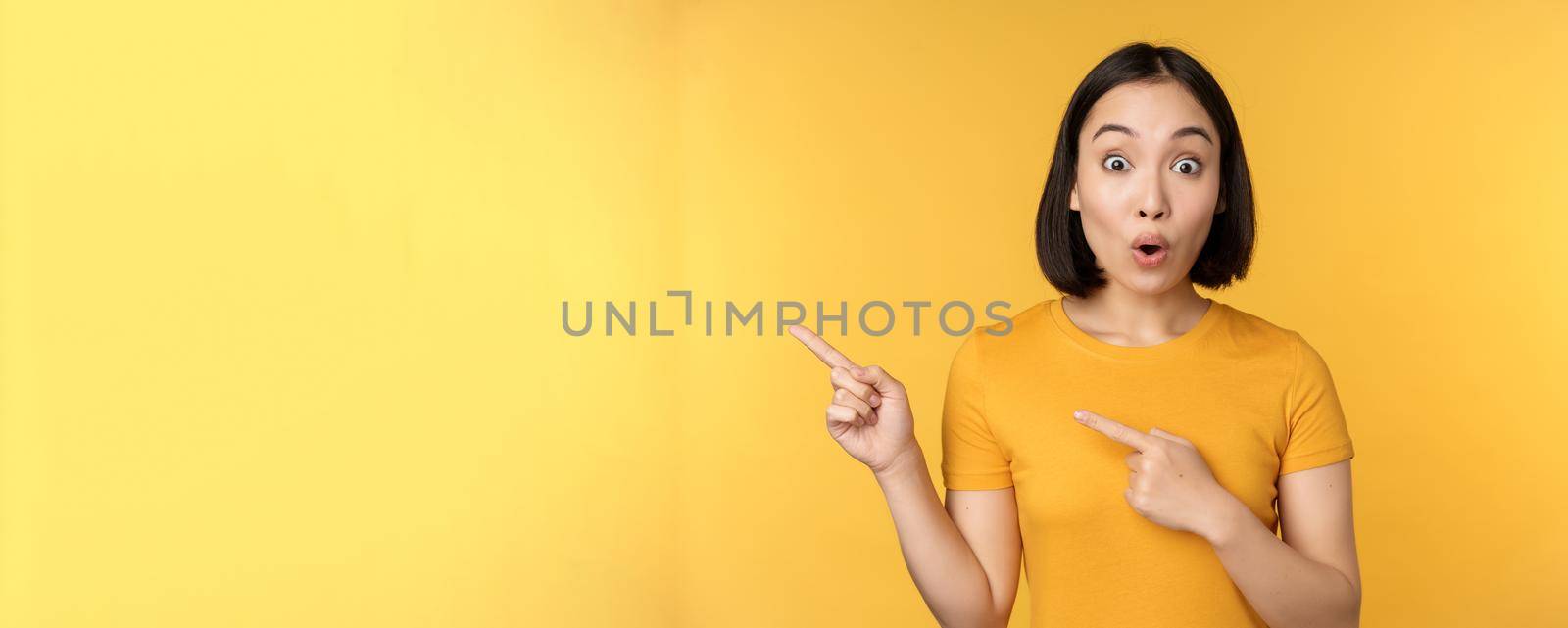 Smiling asian woman pointing fingers left, showing advertisement on empty copy space, standing over yellow background.