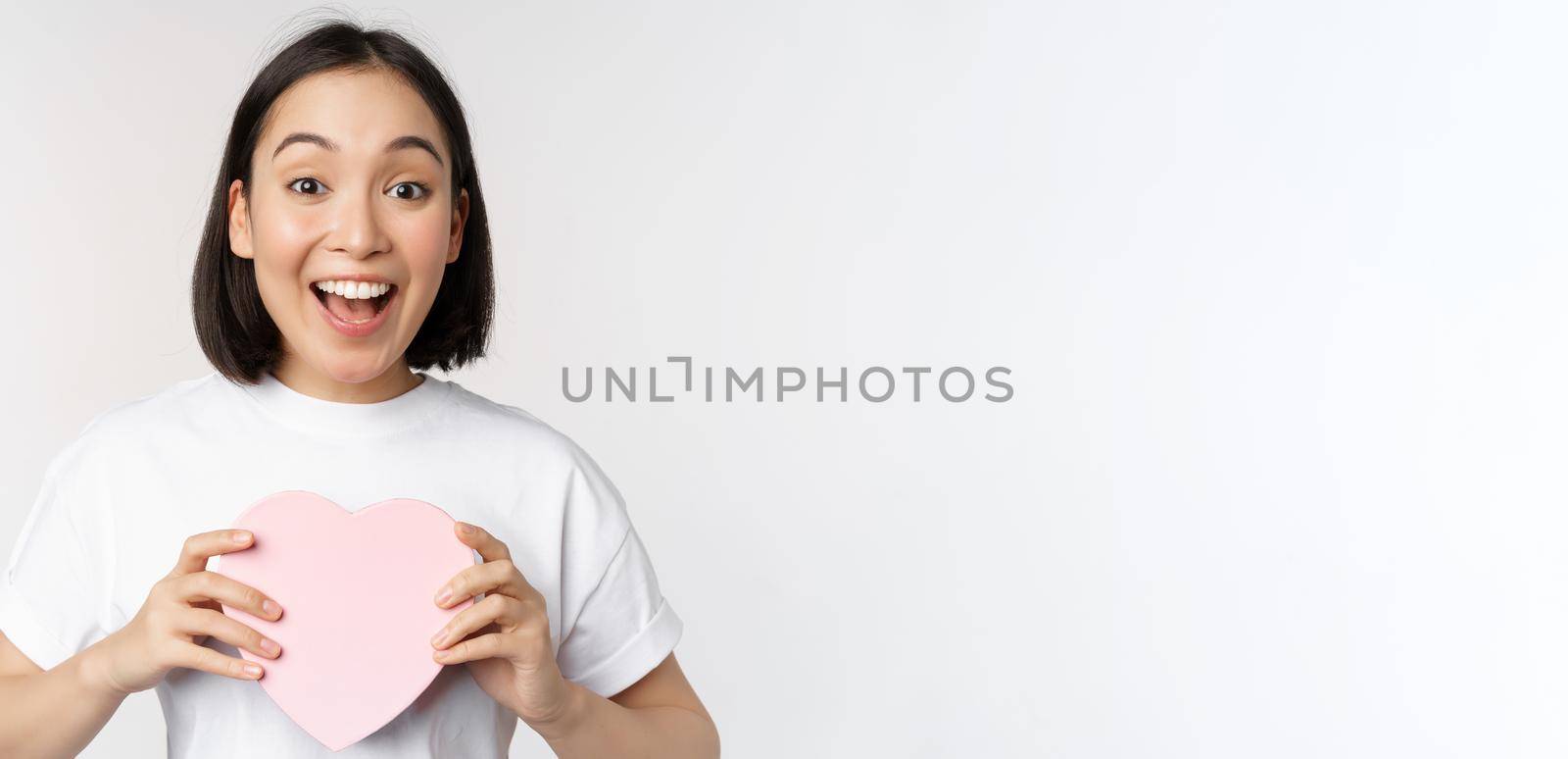 Valentines day. Happy asian girl receive romantic gift, holding heart shaped box and smiling excited, standing over white background.