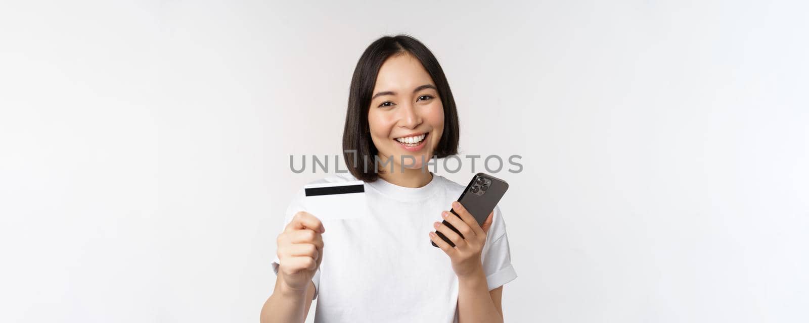 Smiling asian girl using mobile phone, showing credit card, concept of contactless payment, online shopping, standing over white background.