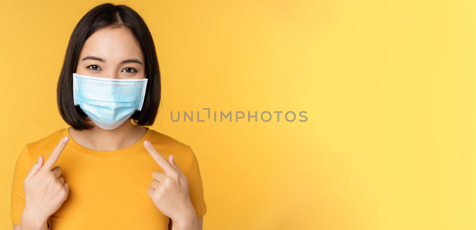 Portrait of smiling asian woman in medical face mask, pointing at her personal protective equipment from covid-19 during pandemic, standing against yellow background by Benzoix
