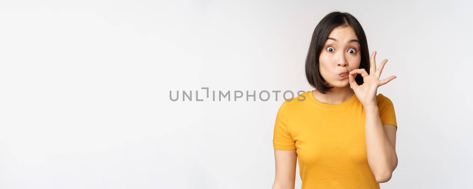 Cute asian girl seal lips, zipping mouth with finger, promise to keep secret, taboo gesture, standing in yellow tshirt over white background.