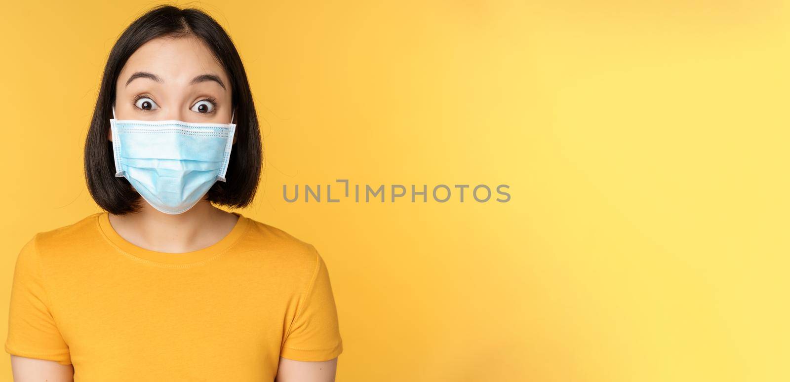 Covid-19 and medical concept. Close up portrait of asian woman in face mask, looking surprised and amazed at news, standing over yellow background by Benzoix