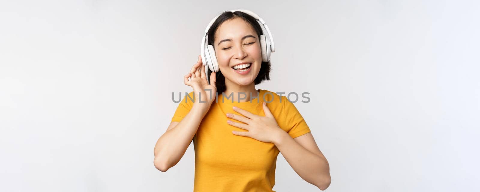 Happy asian girl dancing, listening music on headphones and smiling, standing in yellow tshirt against white background. Copy space