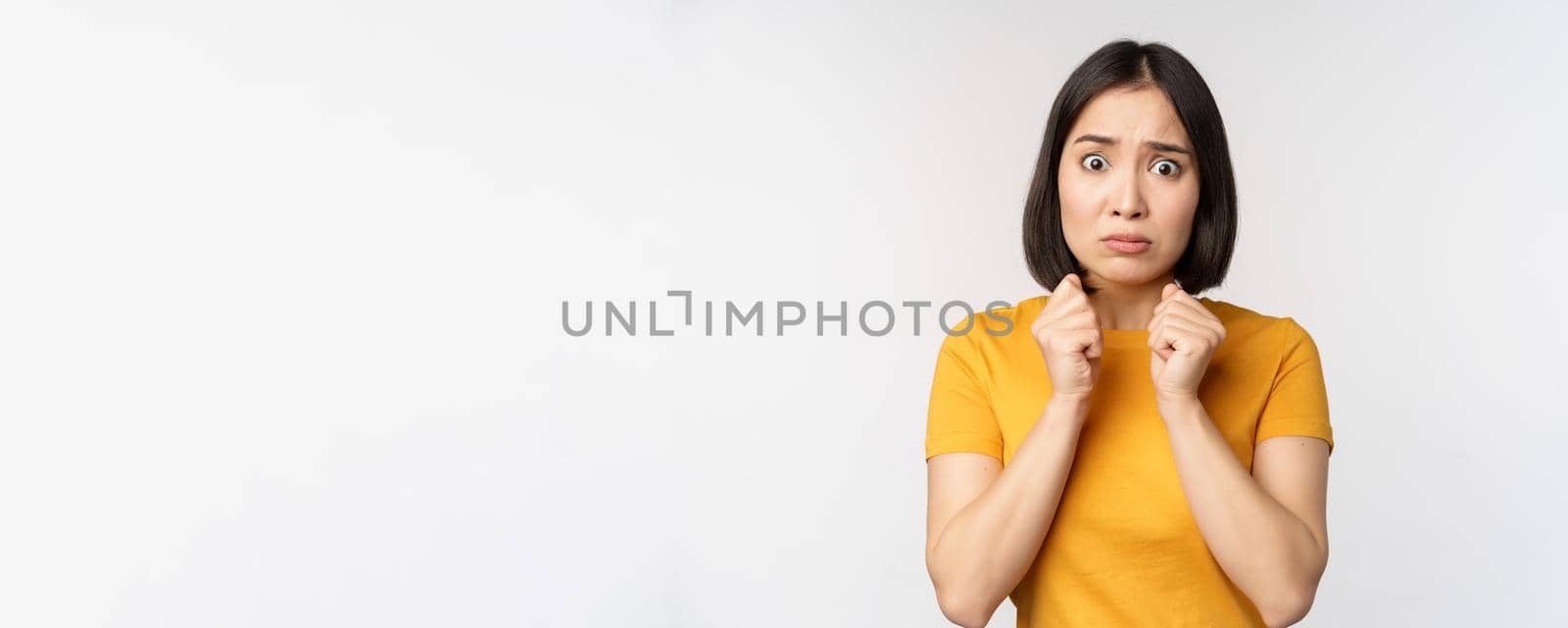 Portrait of scared asian woman shaking from fear, looking terrified and concerned, standing anxious against white background by Benzoix