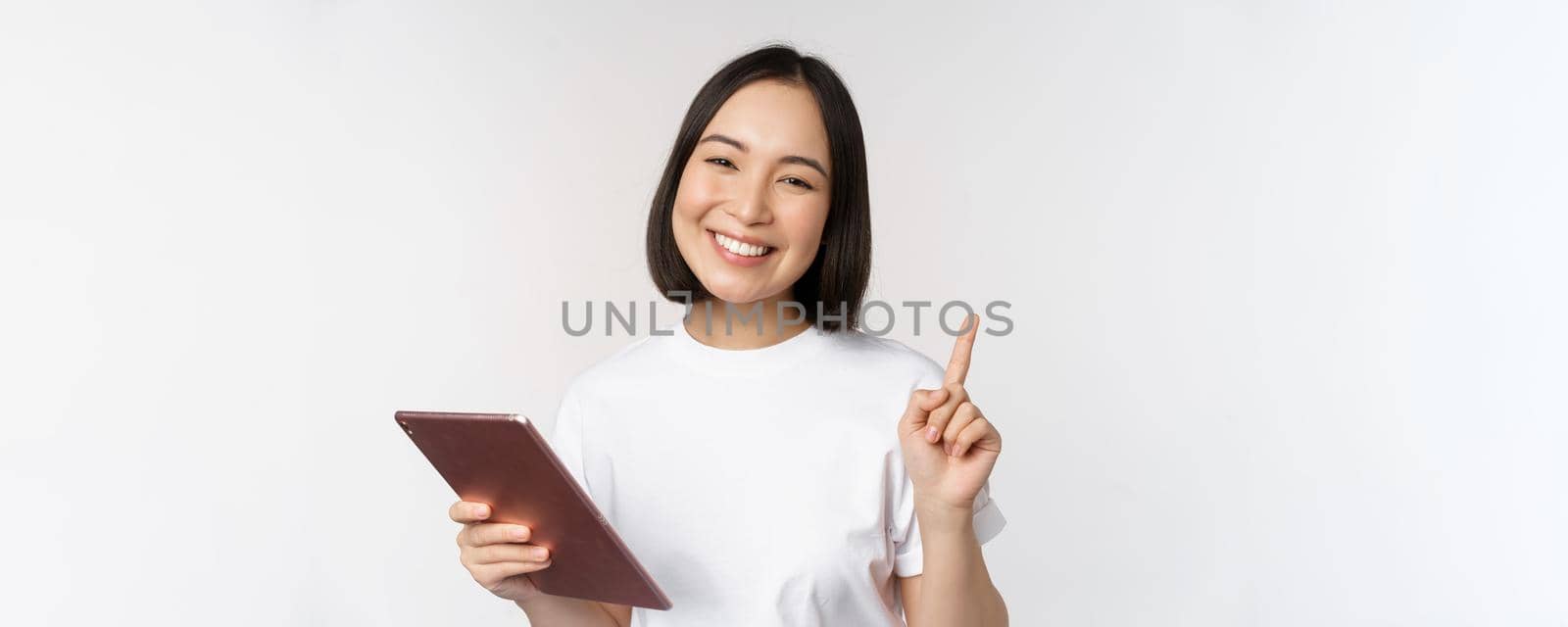 Enthusiastic asian woman with tablet, raising finger and looking amazed, pointing up, standing over white background by Benzoix
