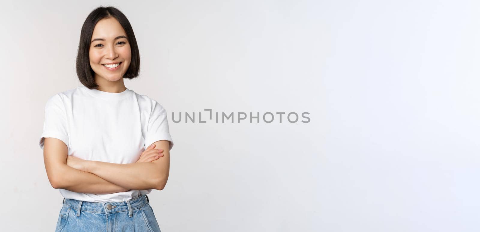 Portrait of happy asian woman smiling, posing confident, cross arms on chest, standing against studio background by Benzoix