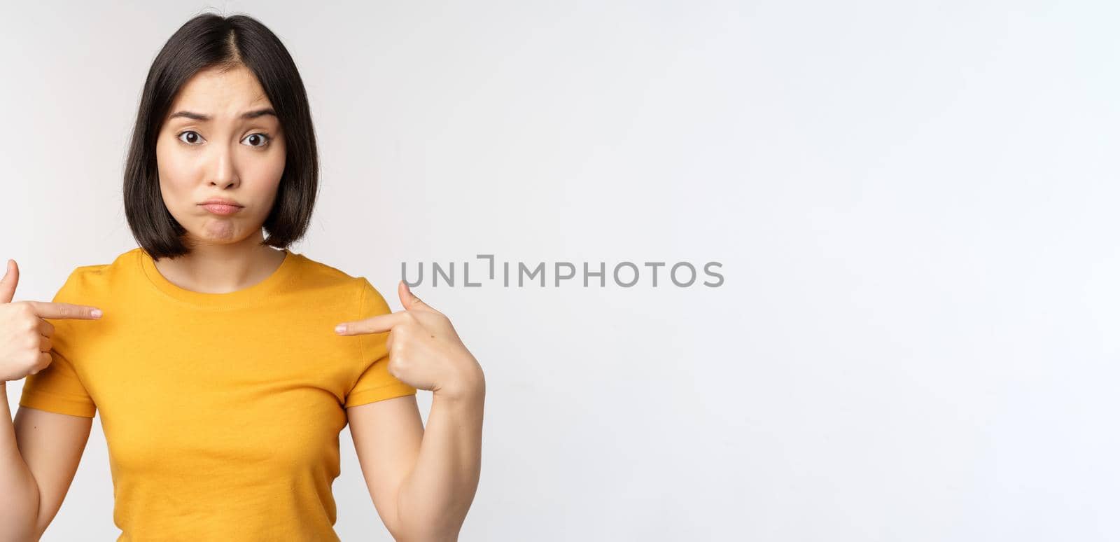 Confused asian woman pointing at herself, looking in disbelief, standing over white background by Benzoix