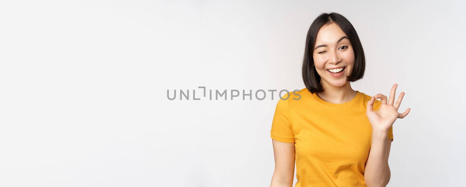 Beautiful young woman showing okay sign, smiling pleased, recommending smth, approve, like product, standing in yellow tshirt over white background by Benzoix