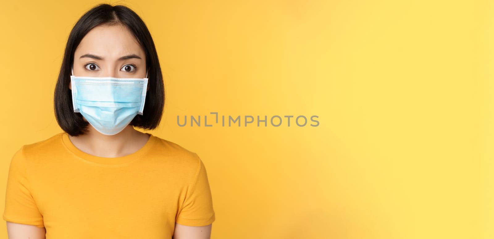 Portrait of shocked asian woman looking concerned and startled at camera, wearing covid-19 medical face mask, standing against yellow background.